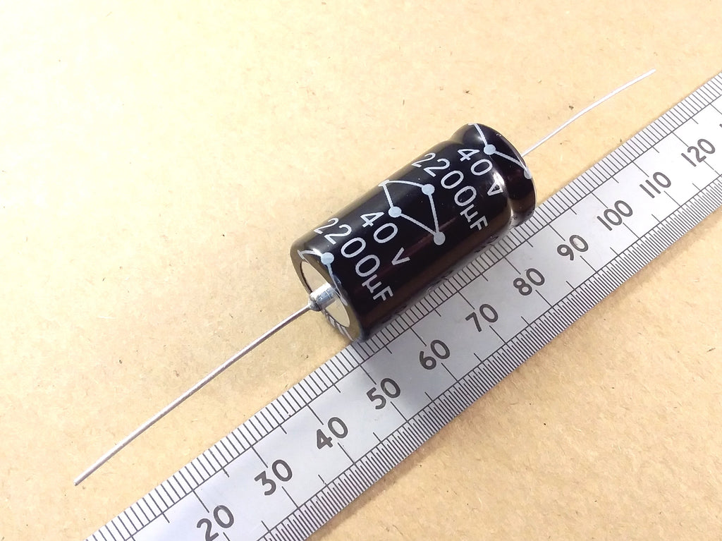 A black cylindrical electrolytic capacitor with white printed markings, shown diagonally next to a metal ruler on a beige background.