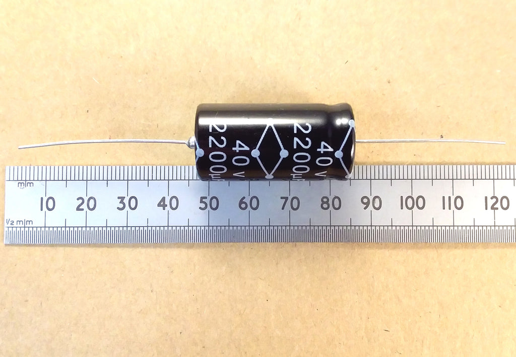 A black cylindrical electronic capacitor with white printed markings, shown horizontally above a metal ruler on a beige background.