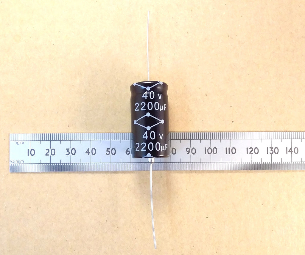 A black cylindrical electrolytic capacitor with white printed markings reading 40 V and 2200 uF, positioned vertically on top of a metal ruler against a tan background.