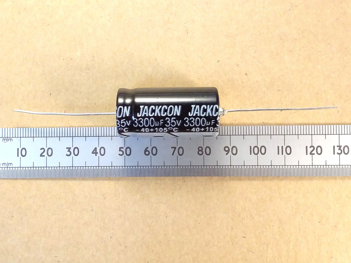 A black axial electrolytic capacitor with white printed text reading 'JACKCON' and '35V 3300uF 35V 3300uF8' is shown horizontally on top of a metal ruler against a tan background.