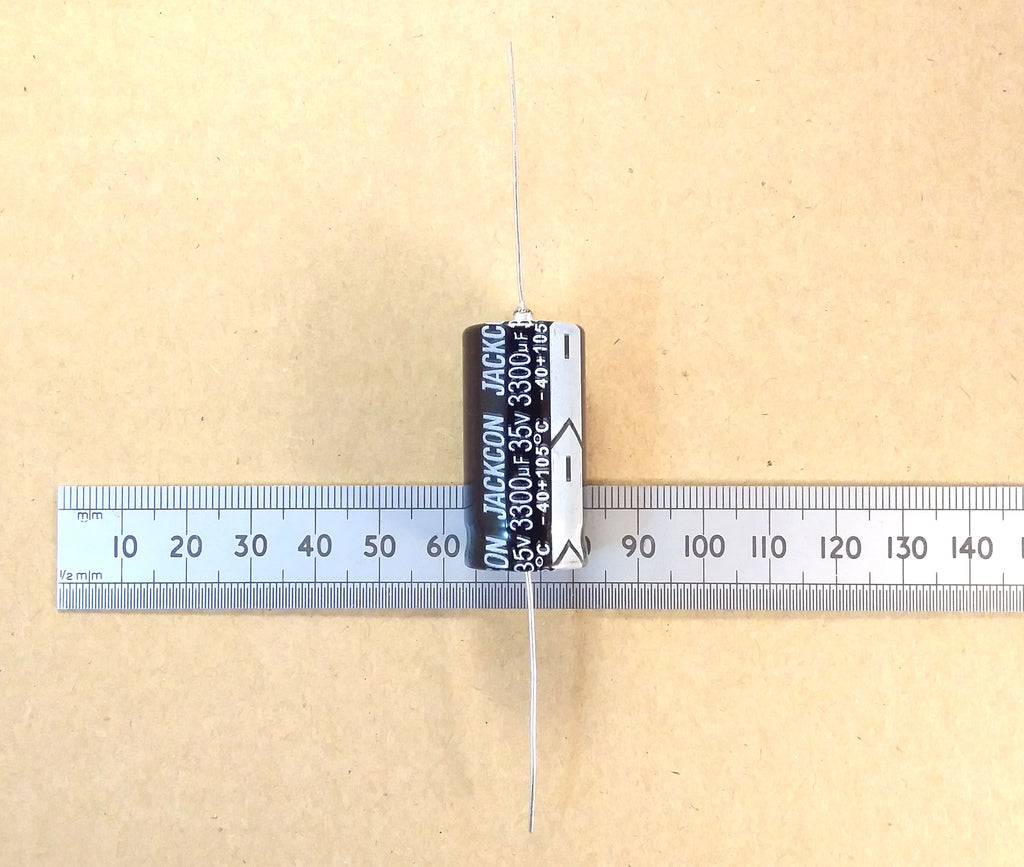 A single cylindrical axial electrolytic capacitor with a black body and white printed text, shown vertically on a metal ruler against a tan background. The capacitor is labeled Jackon 330uF 25V.