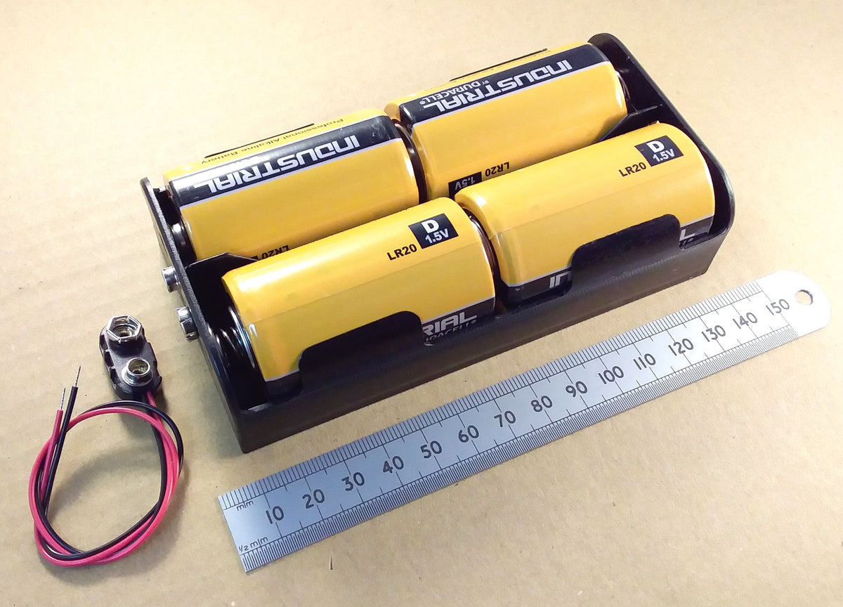 HJW Electronics. A 4-cell battery pack in a black holder on a beige surface, with a metal ruler and a small red/black wire component placed beside it for scale and context.