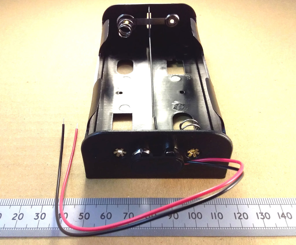 An HJW Electronics black plastic dual battery holder with two exposed wires (red and black) and a metal ruler below for scale, photographed on a beige surface.