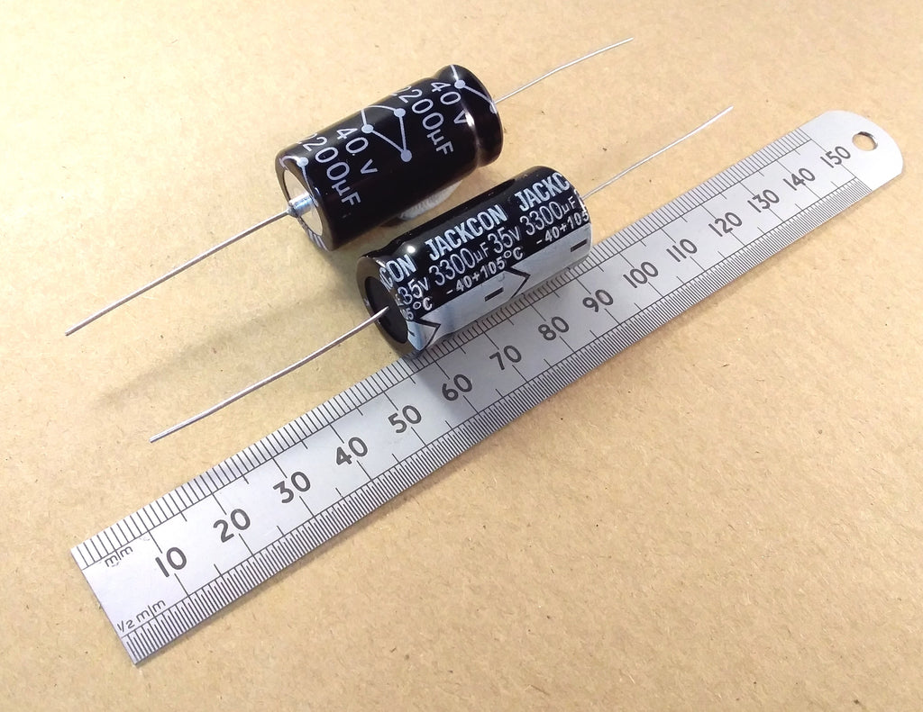Two black cylindrical electrolytic capacitors with silver leads, shown next to a metal ruler on a beige background. The capacitors have white printed specifications including 'JACKSON JONES' and '330uF'.