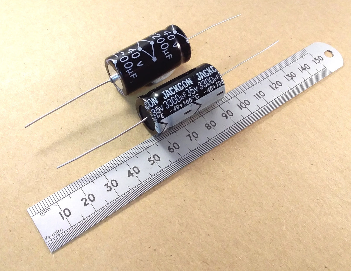 Two black cylindrical electrolytic capacitors with silver leads, shown next to a metal ruler on a beige background. The capacitors have white printed specifications including 'JACKSON JONES' and '330uF'.