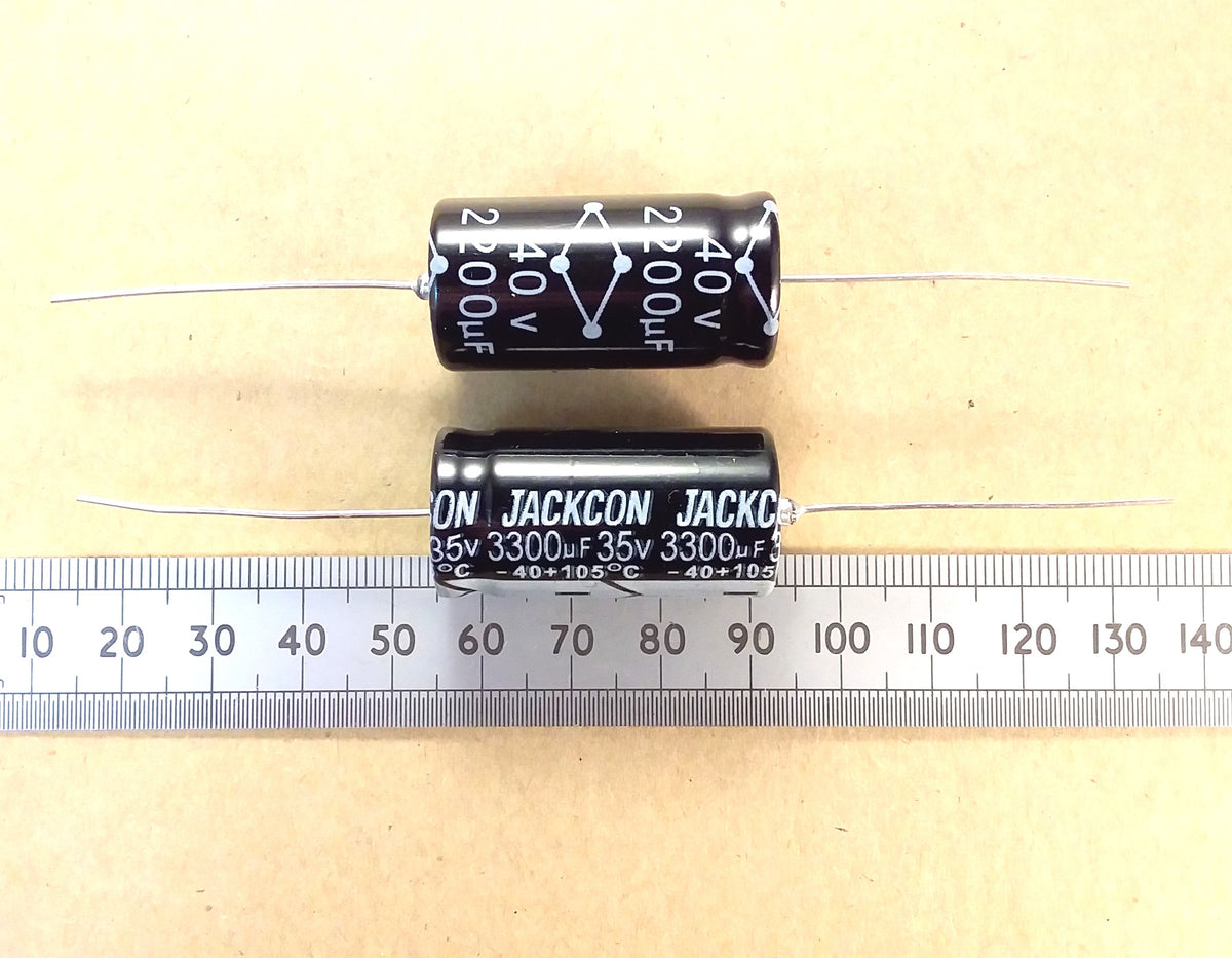 Two black cylindrical capacitors with white printed specifications and metal leads, shown on a beige background with a metal ruler below for scale. The text 'JACKSON' is visible on the capacitors.