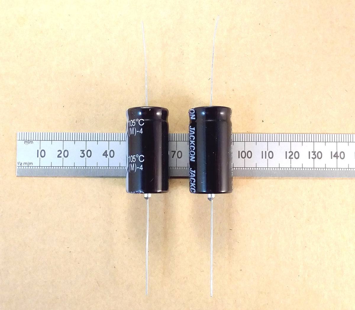 Two black cylindrical electrolytic capacitors with white text, shown side-by-side on a beige background with a metal ruler for scale.